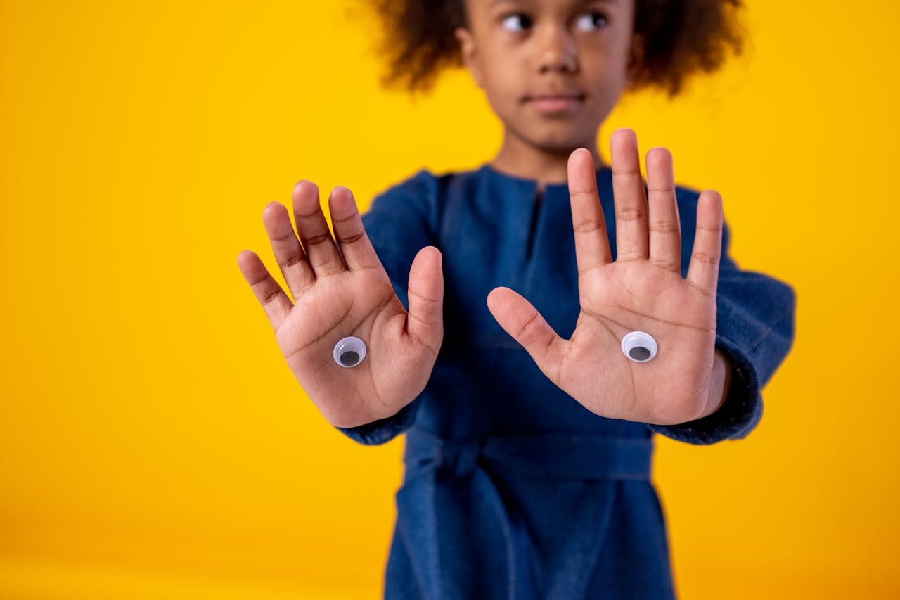 A young girl in a blue dress shows her palms decorated with googly eyes against a bright yellow background.