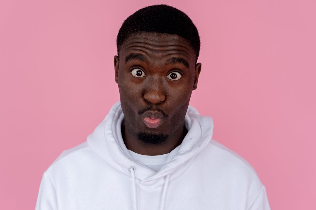 Studio portrait of a young man in a hoodie making a funny face against a pink background.