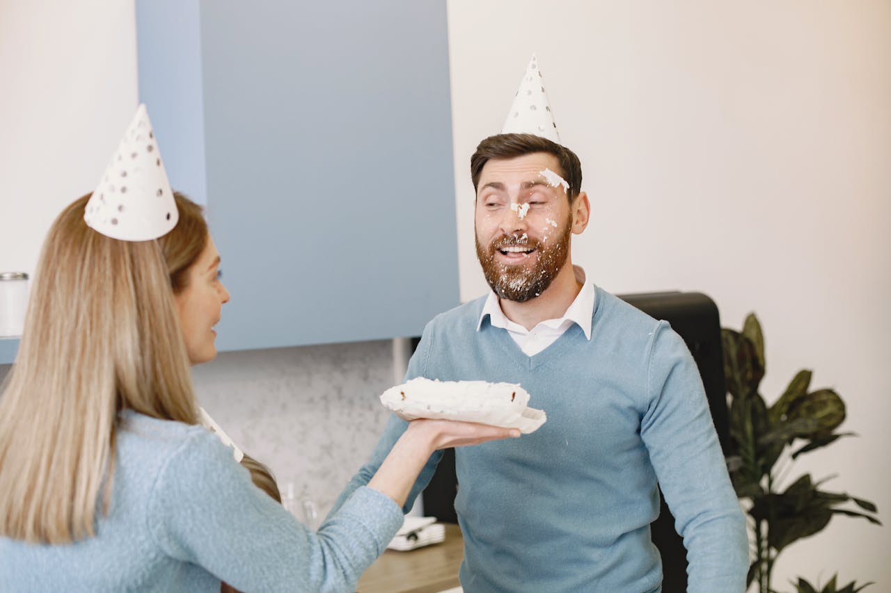 our-story-01 Man and woman sharing a playful cake smash during a birthday celebration indoors.
