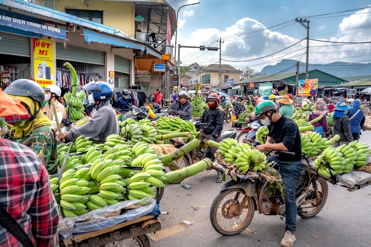 Motorcycle vendors carrying bananas in a bustling outdoor market scene.